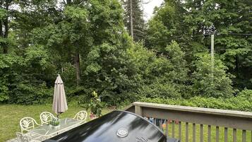 Un barbecue Weber noir sur une terrasse en bois, avec une table et des chaises en verre, le tout entouré d’une végétation luxuriante.