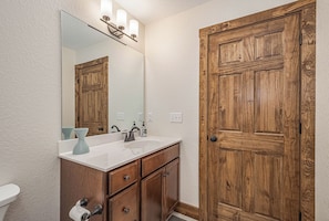 A bathroom with a wooden door, a white sink with a mirror above it, and a light fixture. - Happy Ours by Taylor Made Deep Creek (McHenry)
