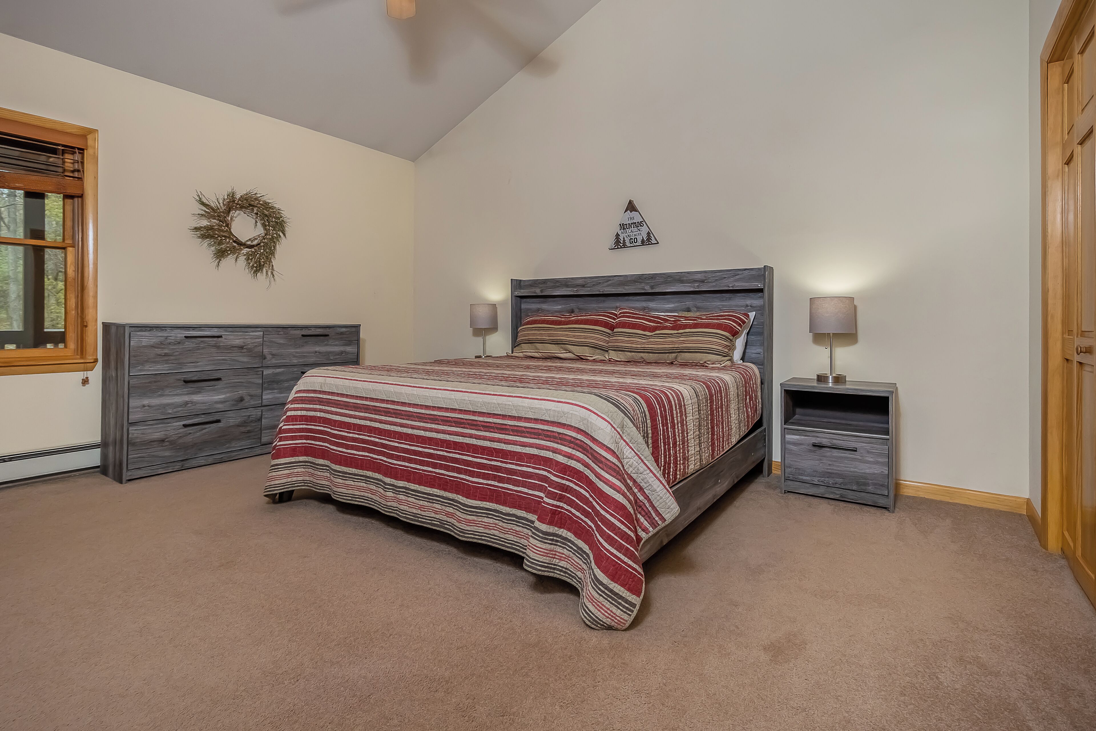 A bedroom with a bed, two nightstands, a wooden headboard, a wall-mounted wreath, and a small wall decoration.