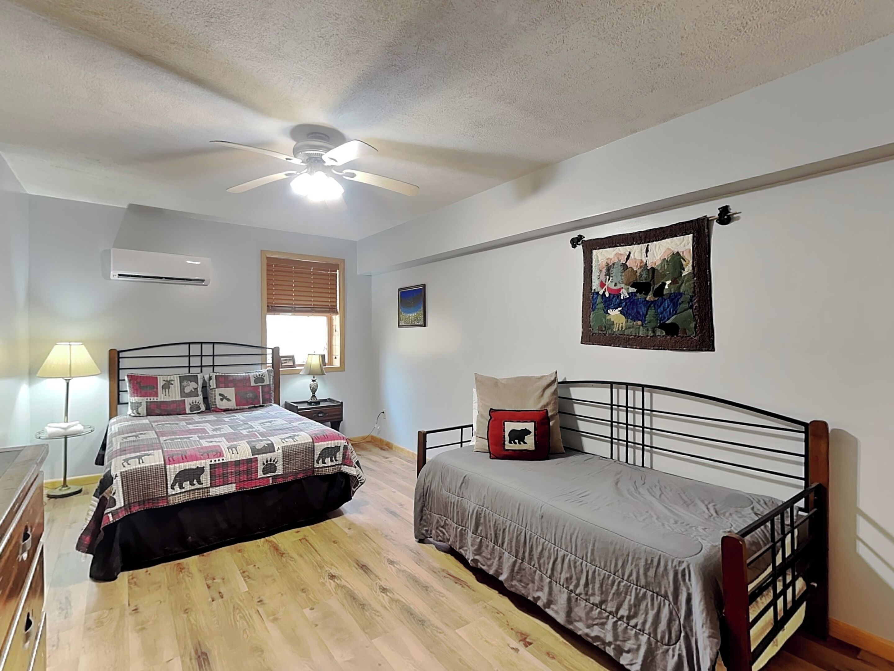 A bedroom with a bed, a bedside table, a lamp, a ceiling fan, a window with blinds, and a wall-mounted painting.