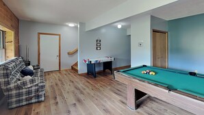 A room with a pool table, a sofa, a small table with pool cues, and a staircase. - Fairway Breeze by Taylor Made Deep Creek (McHenry)