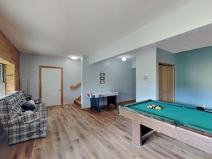 Ein Raum mit Poolbillardtisch, einer Couch, einem kleinen Tisch mit Billardstöcken und einer Treppe.