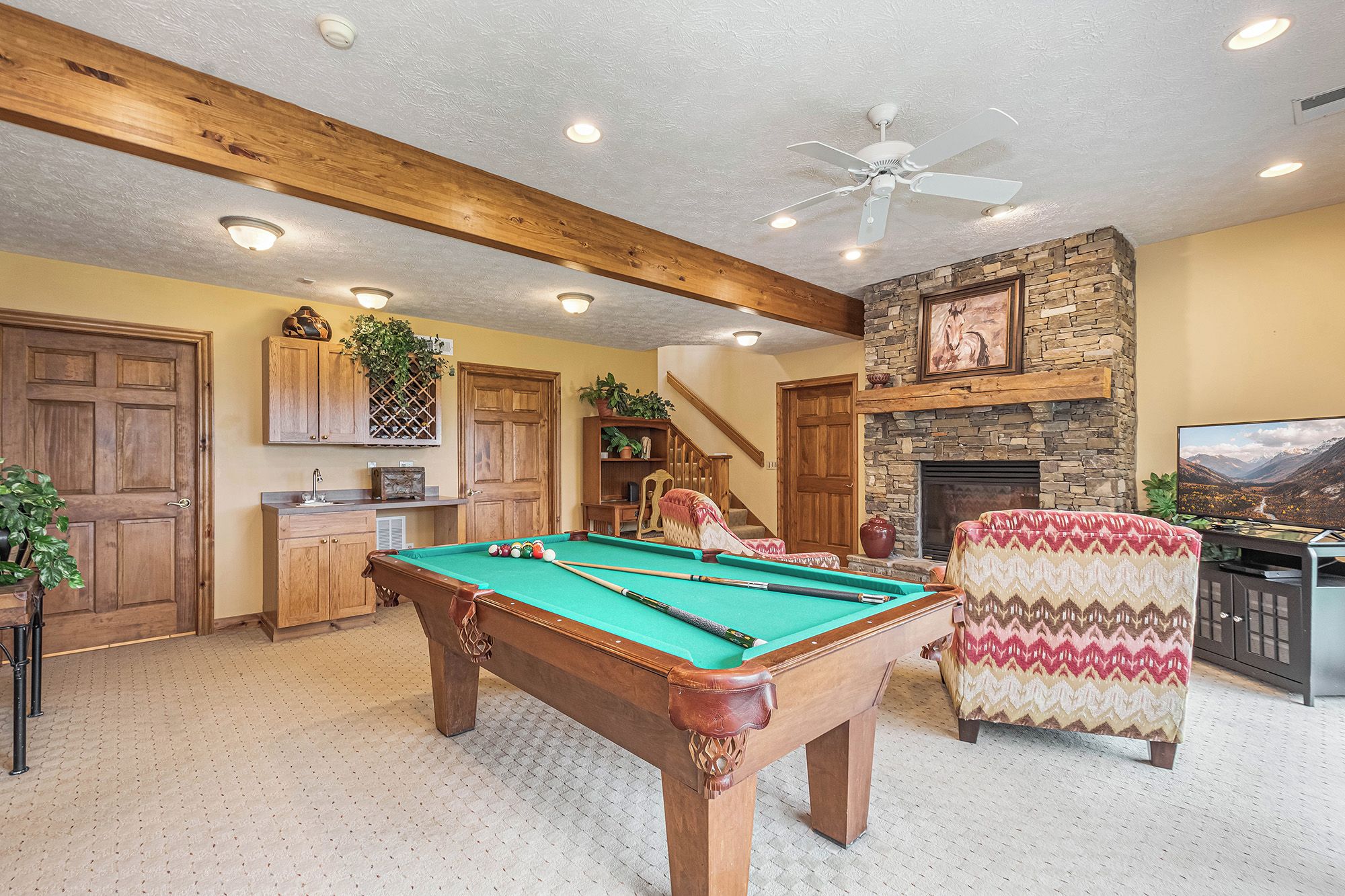 A pool table in a room with a stone fireplace, wooden ceiling beams, and a television on a stand.
