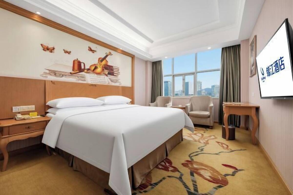 A hotel room with a large bed, a desk, a chair, a TV, and a window with a city view.