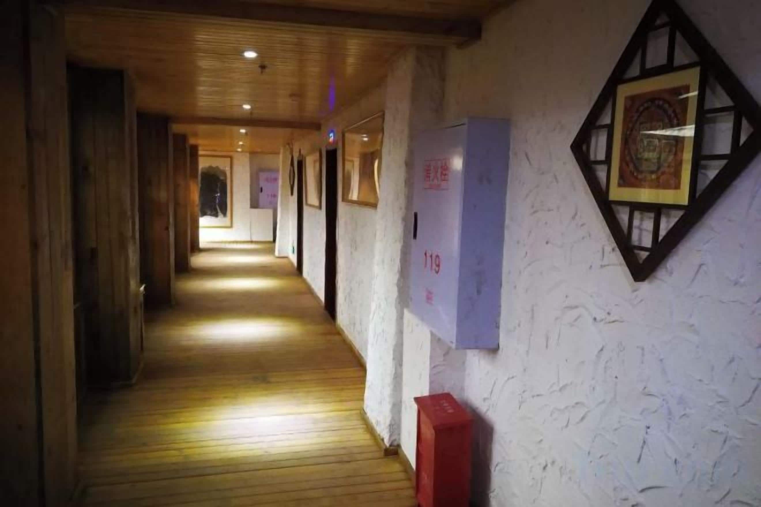 A corridor with wooden flooring, framed pictures on the walls, and a red trash bin.