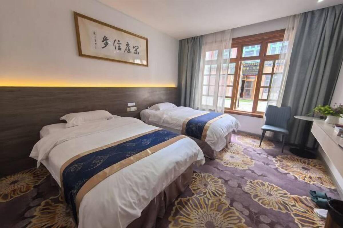 Een hotelkamer met twee bedden, een bureau en een stoel. Er is een raam met gordijnen en een foto in een lijst aan de muur.