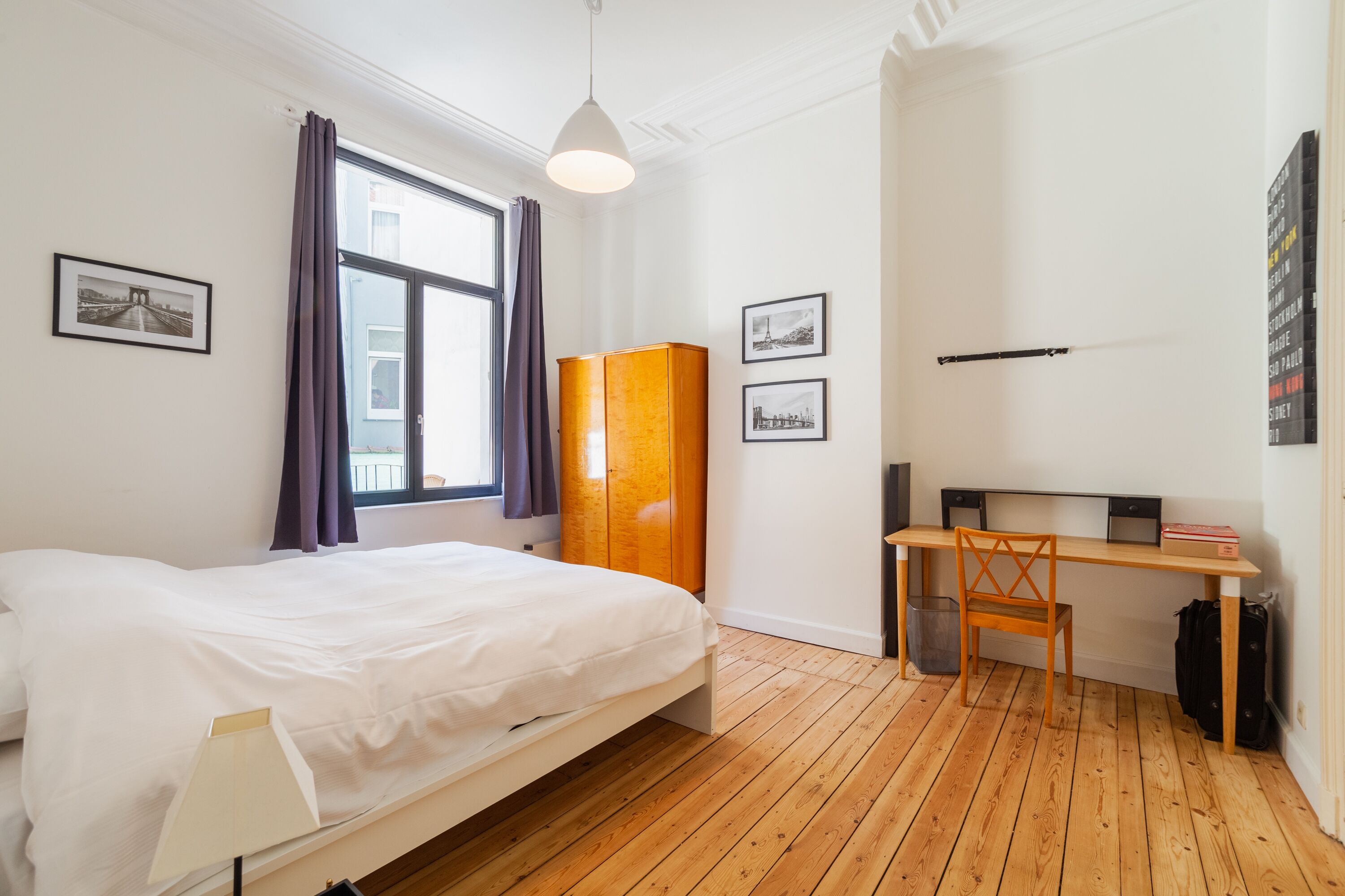 A bedroom with a bed, a desk, and a chair. There are framed pictures on the wall.