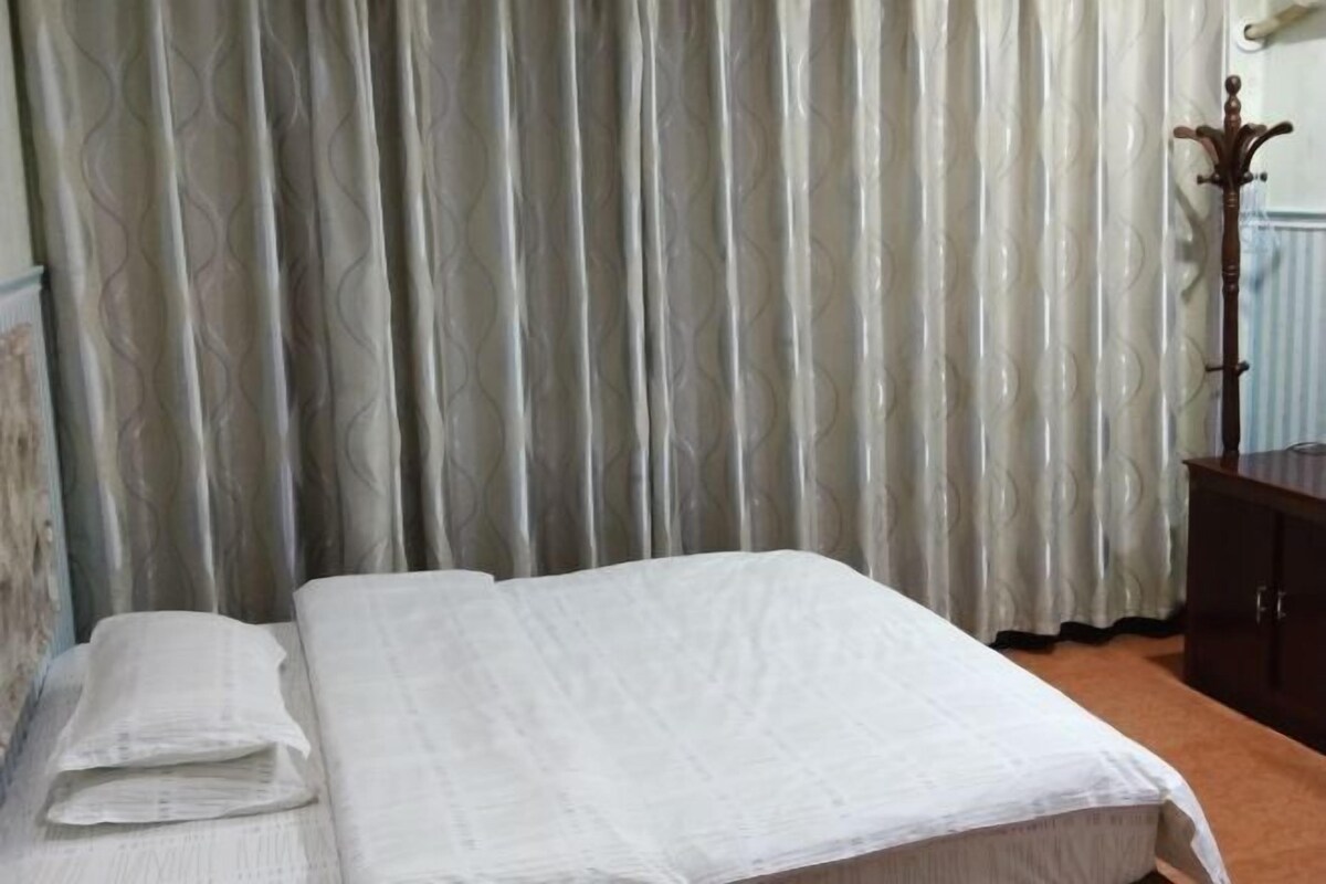 Cama bem arrumada com colcha branca e travesseiros, mesa de cabeceira de madeira com abajur e cortina estampada ao fundo.