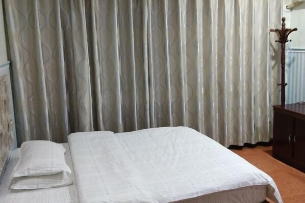 Cama bem arrumada com colcha branca e travesseiros, mesa de cabeceira de madeira com abajur e cortina estampada ao fundo.