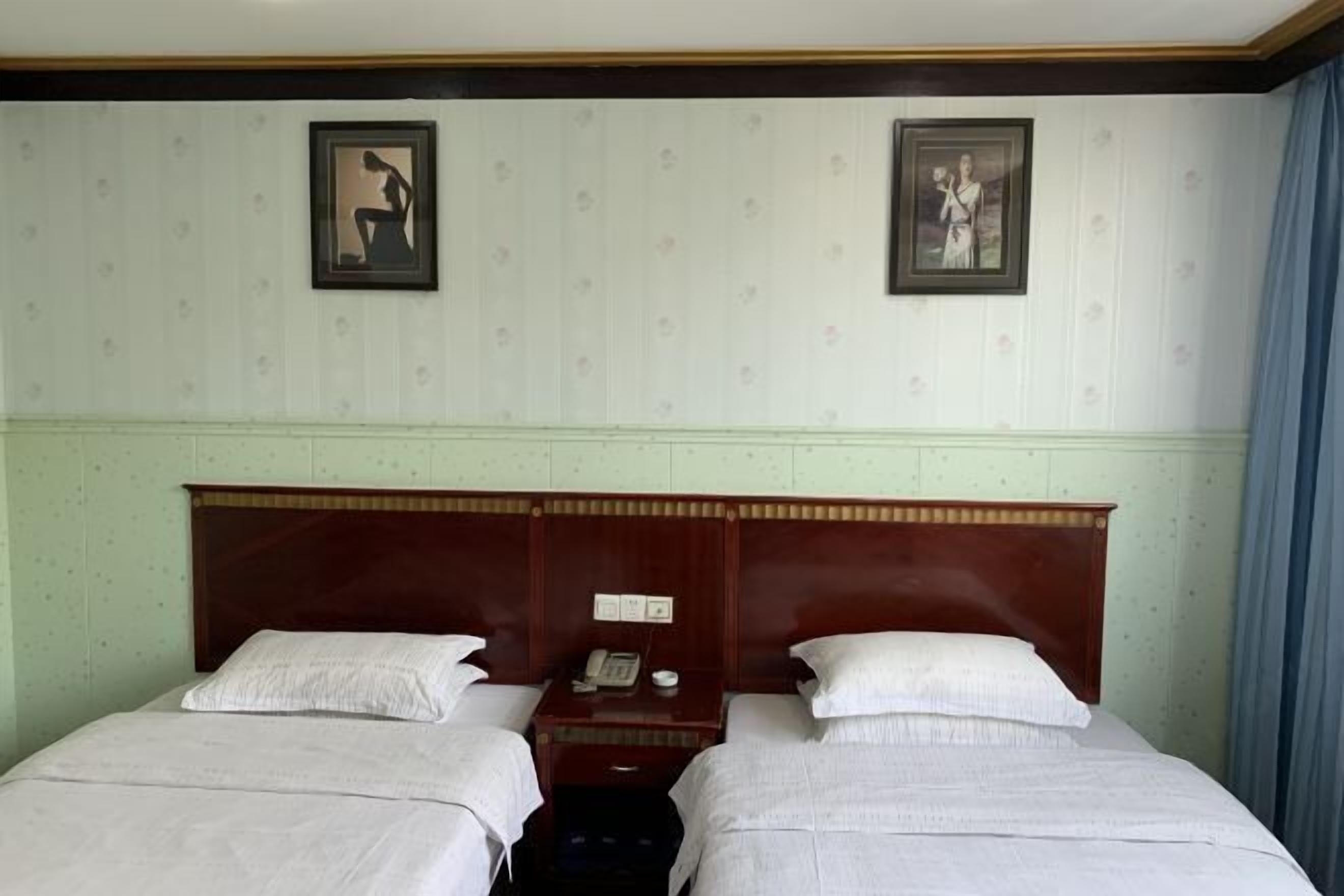 Ein Hotelzimmer mit zwei Einzelbetten, einem Holzkopfende, einem Nachttisch mit Telefon und Lampe sowie zwei gerahmten Bildern an der Wand.