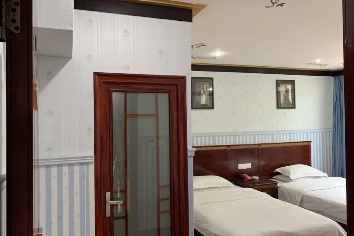 Quarto de hotel com duas camas de solteiro, cabeceira de madeira, mesinha de cabeceira e porta com painel de vidro.