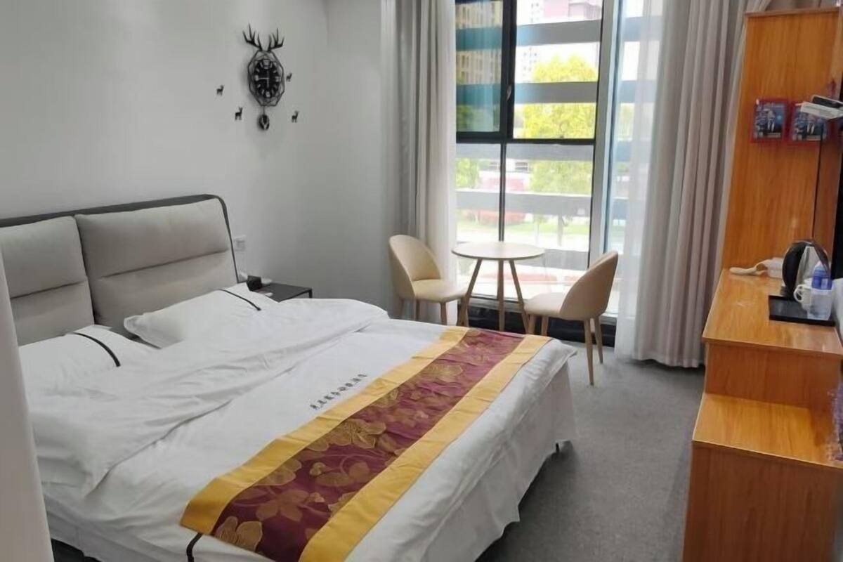 Habitación de hotel con cama, una mesita, dos sillas, un escritorio de madera y un ventanal con cortinas.