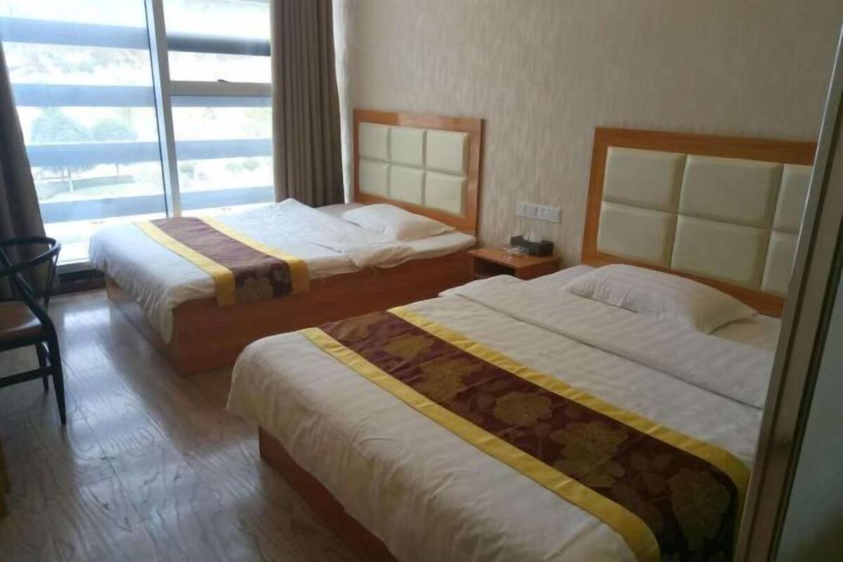 Habitación de hotel con dos camas, suelo de madera, ventana con cortinas y una silla.