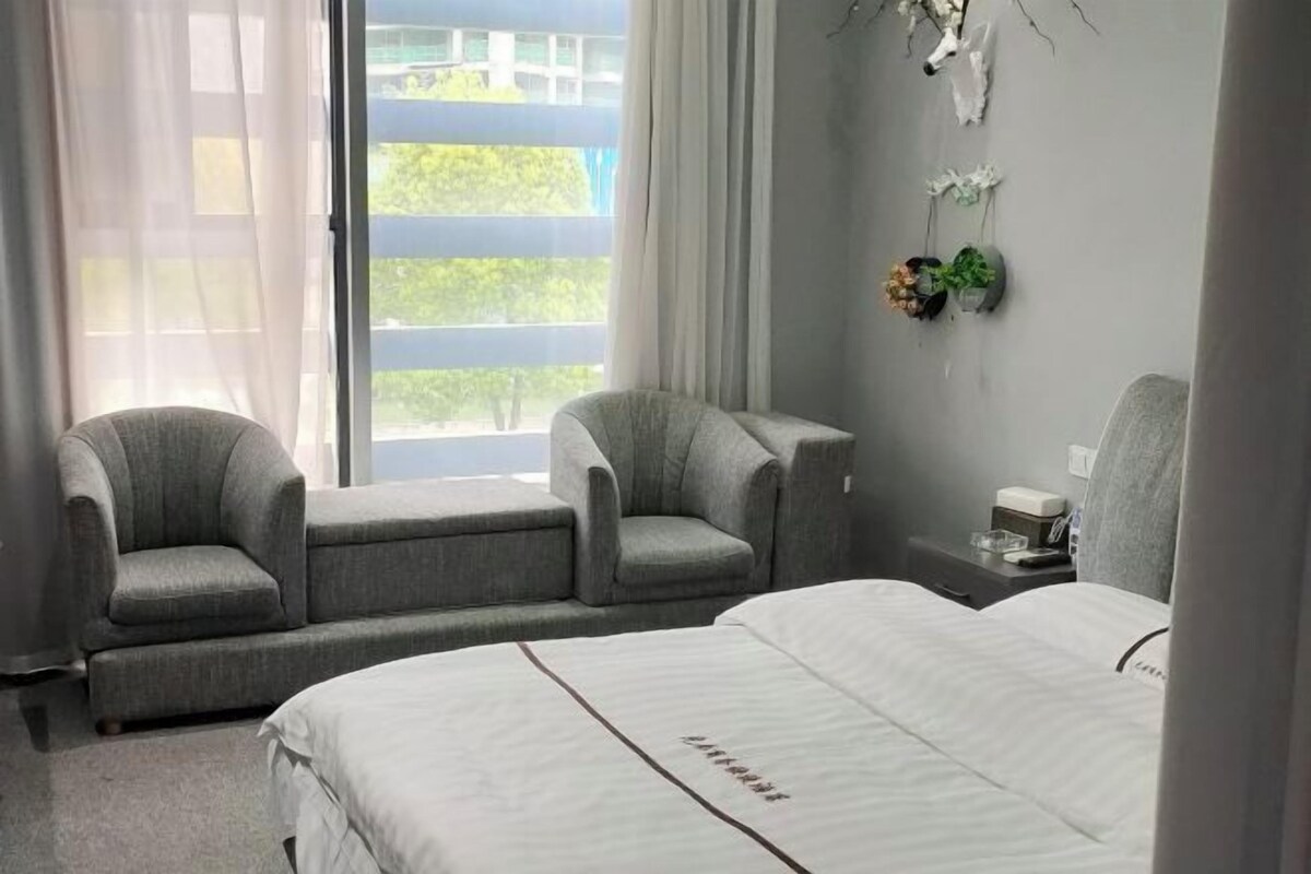 A hotel room with a bed, two armchairs, a nightstand, a window with curtains, and a wall-mounted plant.