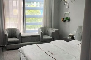 A hotel room with a bed, two armchairs, a nightstand, a window with curtains, and a wall-mounted plant.