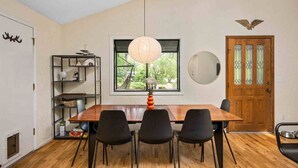 Un coin repas comprenant une table en bois, quatre chaises noires, un luminaire suspendu, une fenêtre donnant sur un espace verdoyant, une porte en bois avec des panneaux de verre et une étagère murale sur laquelle sont disposés divers objets.