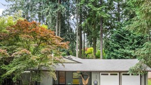 Une maison de plain-pied avec un porche à l’avant, un garage et une cour entourée de grands arbres.