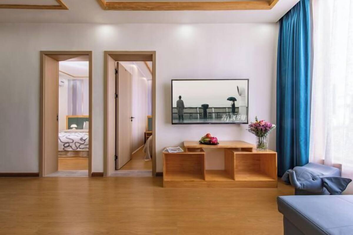A modern living room with a wooden bench, a flat-screen TV mounted on the wall, and a large window with blue curtains.