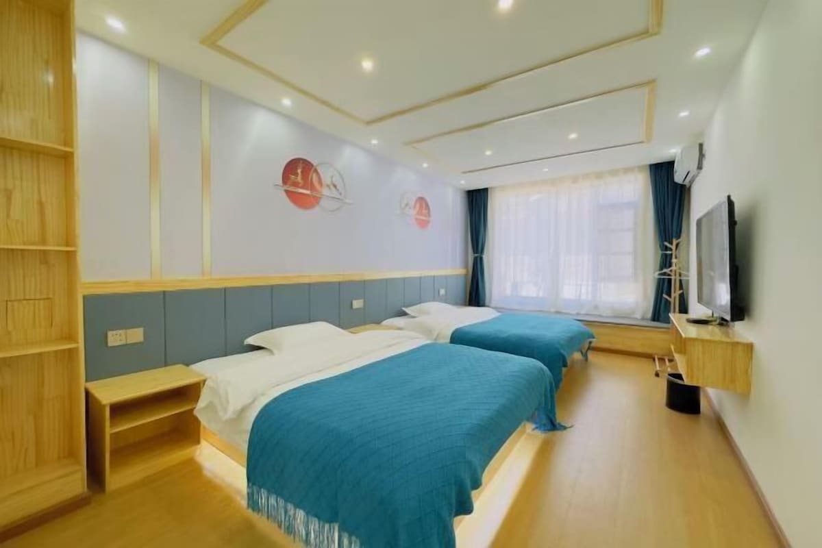 A hotel room with two beds, a TV, a wooden shelf, and a window with curtains.