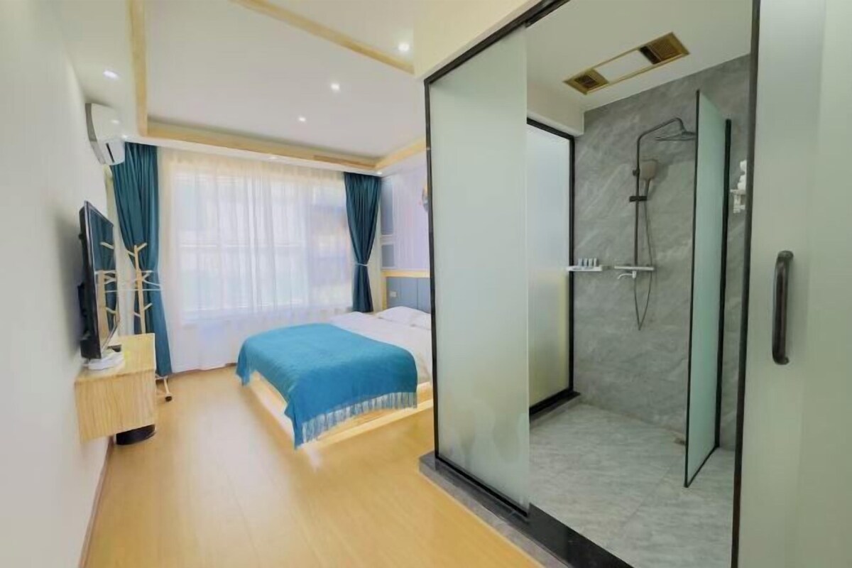A modern hotel room with a bed, a glass-enclosed shower, a TV, and a desk with a lamp.