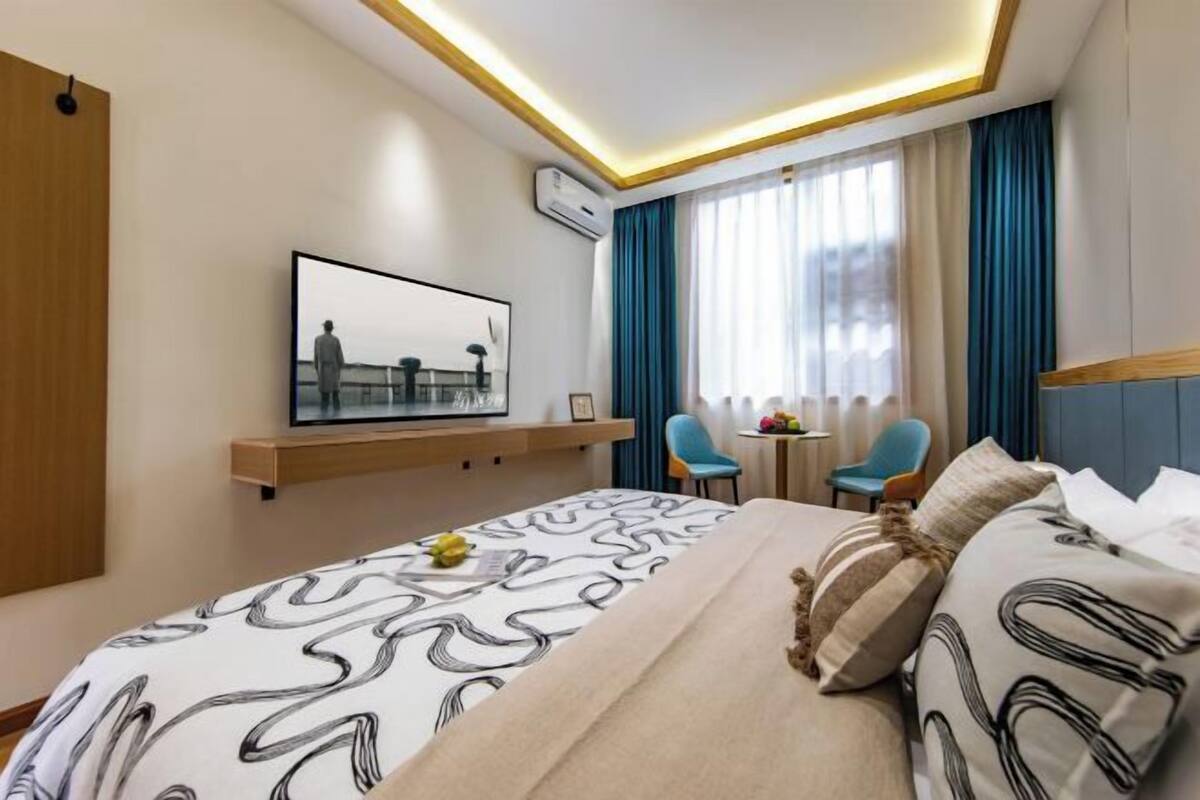 A modern hotel room with a large bed, a mounted TV, a desk with chairs, and a window with curtains.