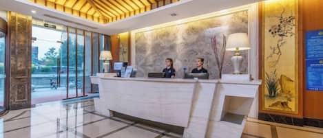 A hotel reception area with a check-in counter, two receptionists, and a large window offering a view of the outdoors.