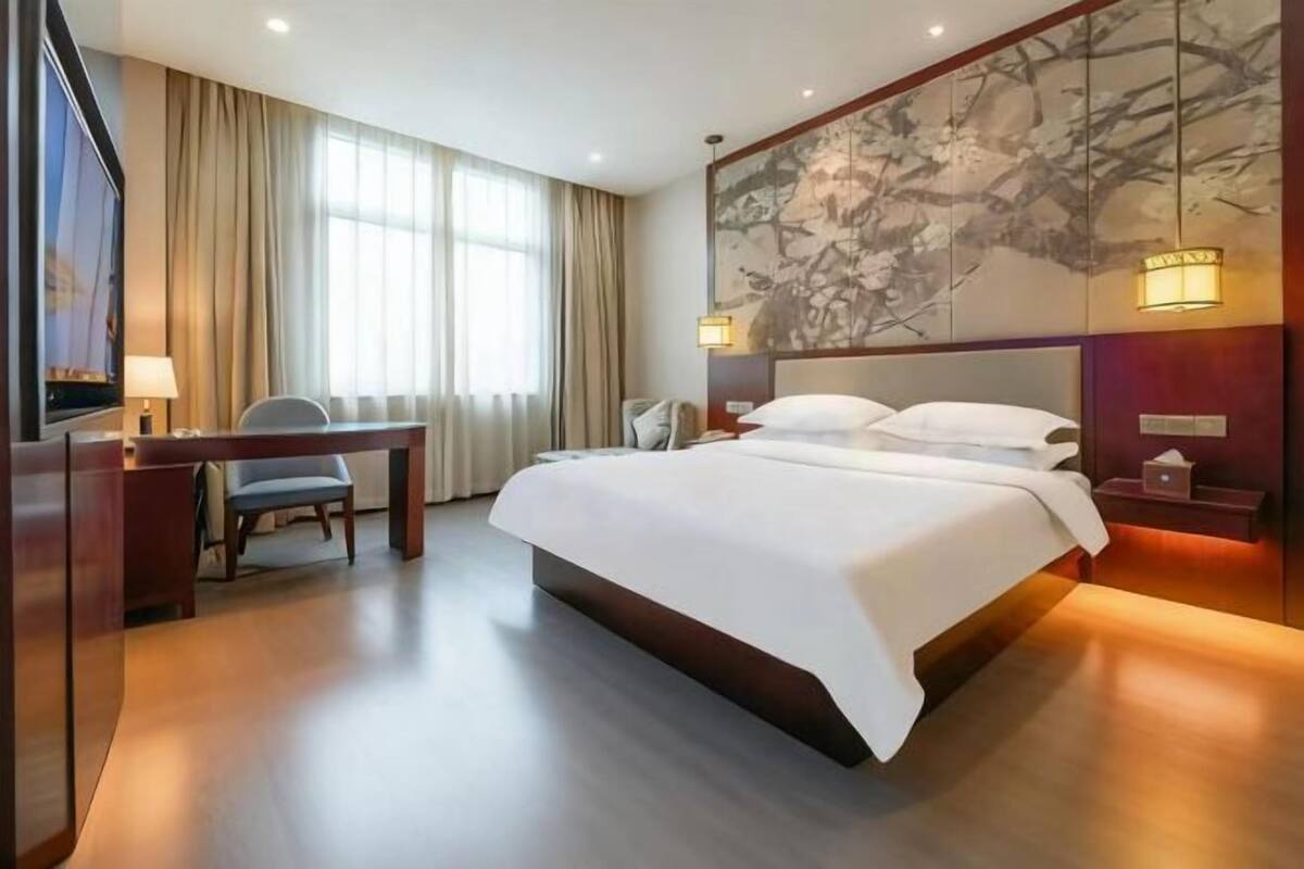 Une chambre d’hôtel avec un grand lit, un bureau avec une chaise, une télévision et une grande fenêtre avec des rideaux.