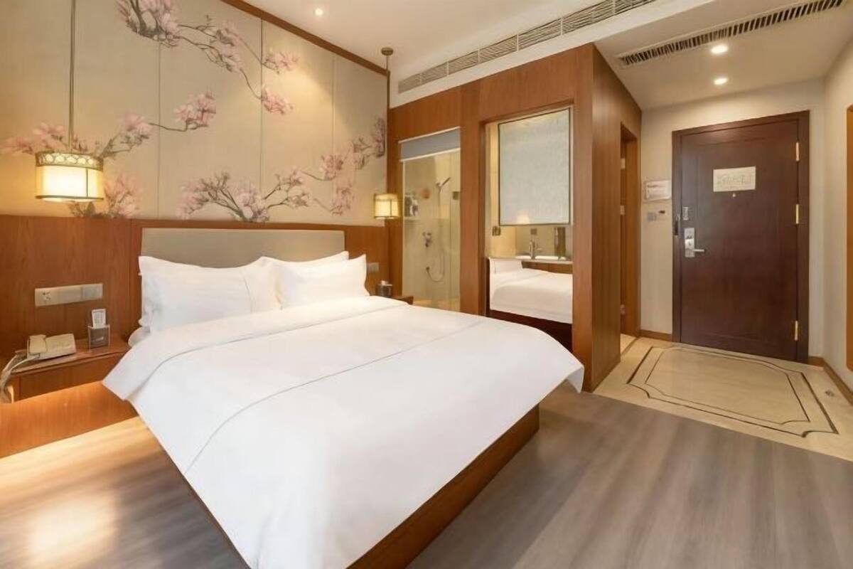 Une chambre d’hôtel avec un grand lit, une tête de lit en bois, une table de chevet avec un téléphone, une lampe fixée au mur, un miroir et une porte avec une pancarte.