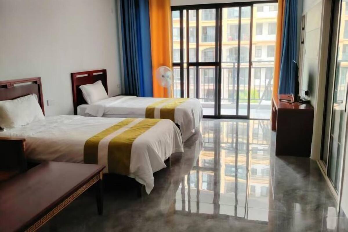 A hotel room with two beds, a bench, a fan, and a large window with a view of buildings.