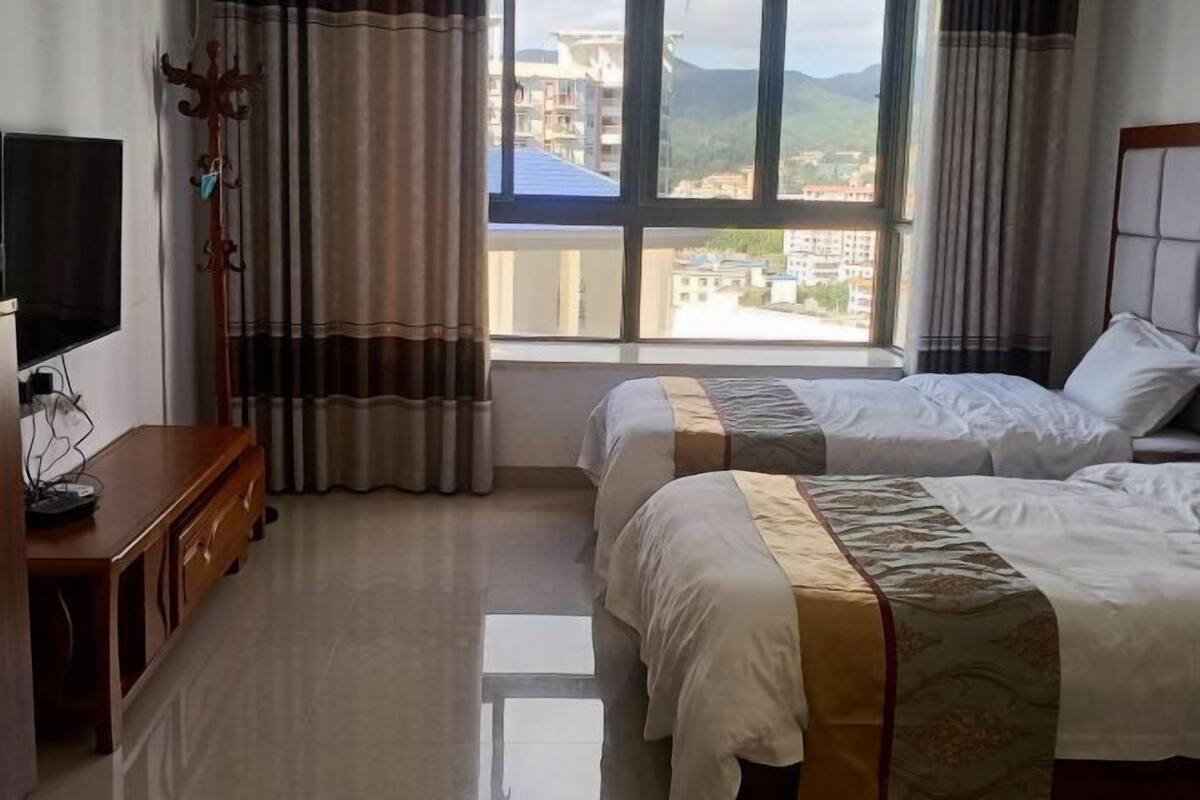 A hotel room with two beds, a TV, a wooden dresser, and a view of buildings and mountains through large windows.