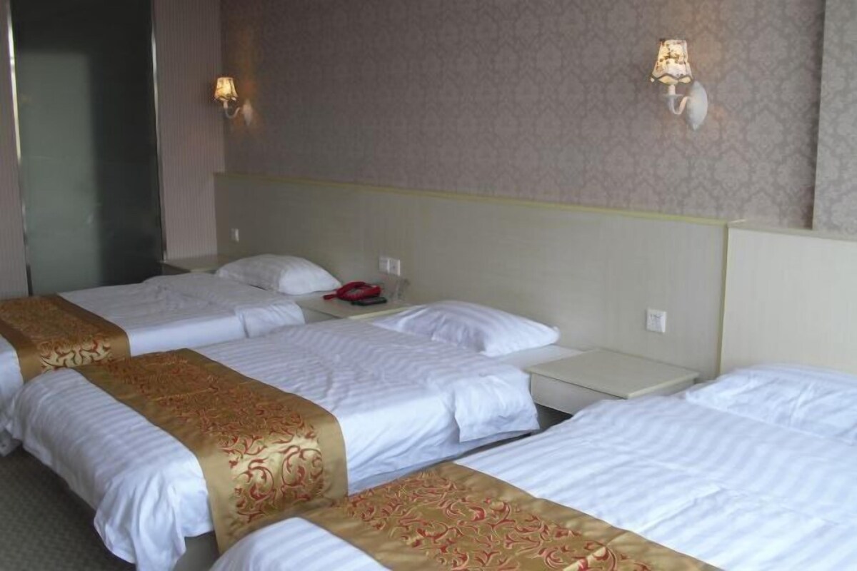 A hotel room with three single beds, each with a white bedspread and a gold decorative runner. There are two bedside tables, one with a lamp and the other with a phone. The walls are adorned with patterned wallpaper.