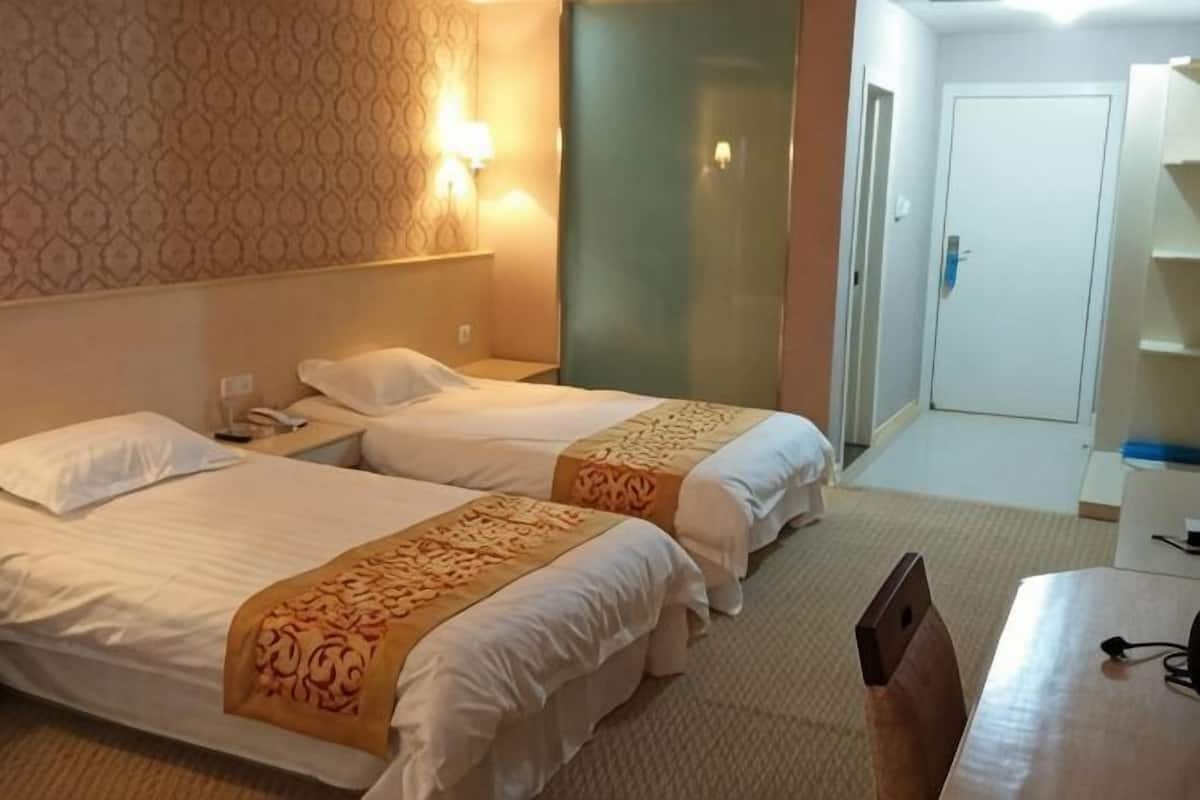 A hotel room with two beds, a desk, a chair, a nightstand, and a wall-mounted lamp.