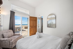 A hotel room with a bed, a sofa, a large window with a view of the beach, and a door leading outside.