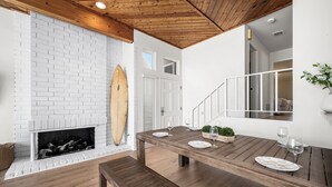 Un coin repas avec une table en bois dressée pour quatre personnes, une cheminée en briques blanches et une planche de surf appuyée contre le mur.