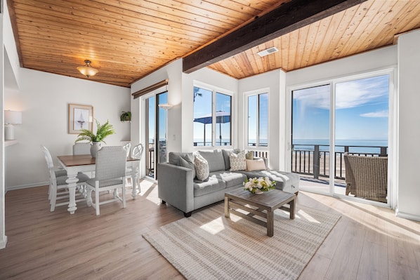 A spacious living area with a wooden ceiling, large windows offering a view of the ocean, a grey sofa, and a dining table with chairs.