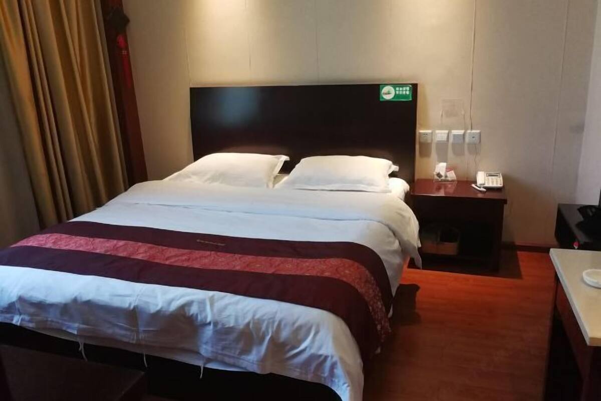 A hotel room with a bed, a nightstand with a phone and a lamp, and a wooden desk.