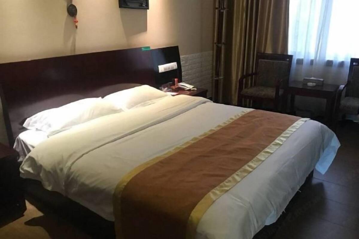 A hotel room with a large bed, a wooden headboard, a bedside table with a lamp, a chair, and a window with curtains.
