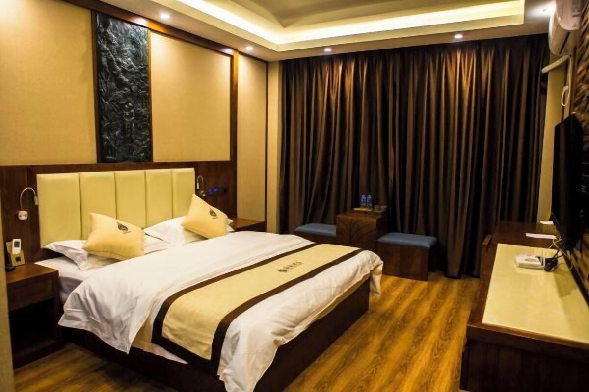 A hotel room with a large bed, a wooden headboard, a desk with a chair, a small table with a chair, a wall-mounted TV, and a curtain.