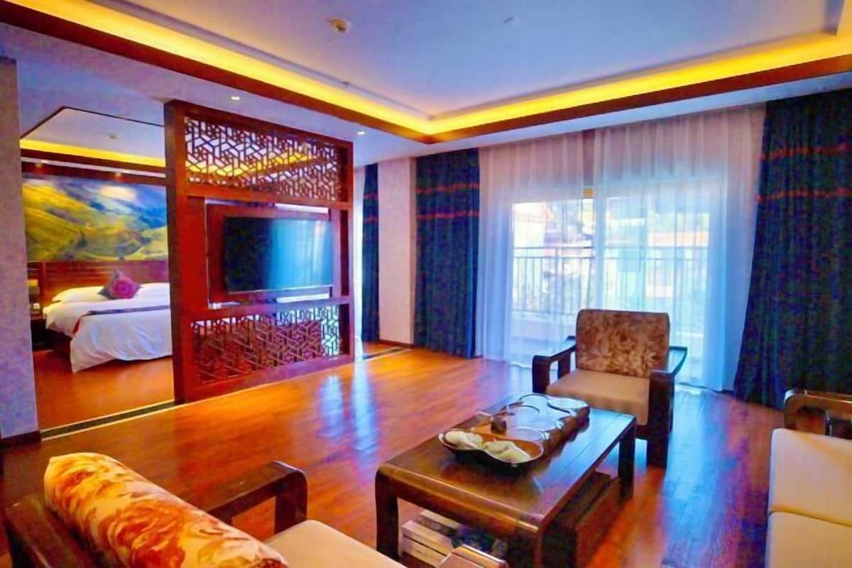 A hotel room with a wooden floor, a sofa, a chair, a TV, and a bed with a painting on the wall.