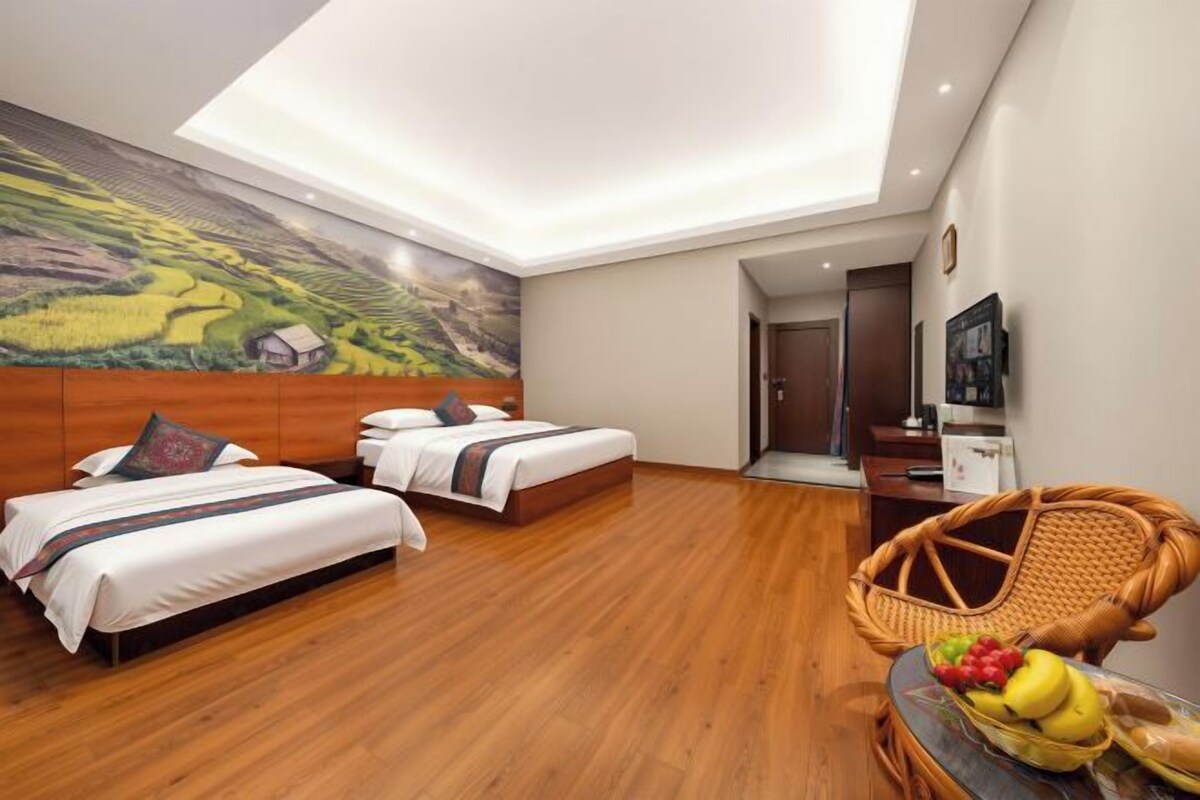 Habitación de hotel con dos camas, suelo de madera, luz de techo, televisión montada en la pared y una silla de mimbre con fruta.