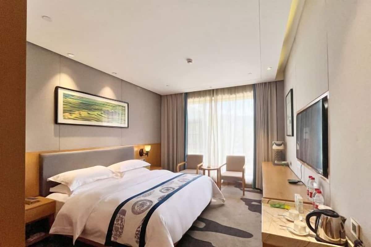 Ein Hotelzimmer mit einem großen Bett, einem Schreibtisch mit Wasserkocher, einem Fernseher und einem Fenster mit Vorhängen.