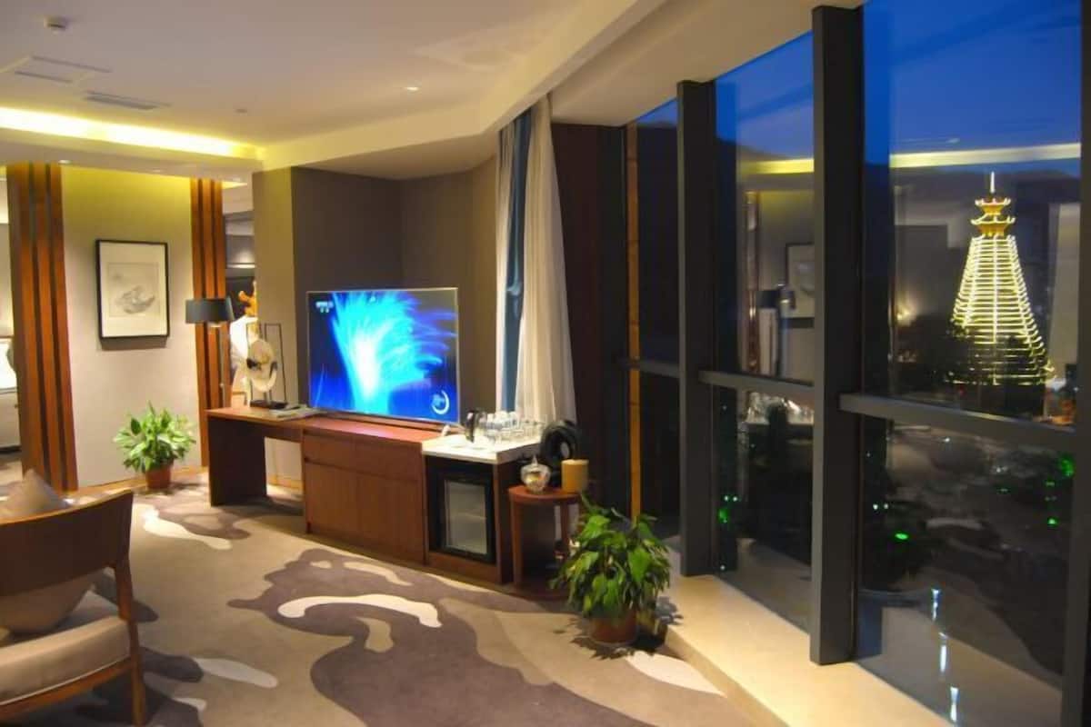 Ein modernes Hotelzimmer mit großem Flachbildfernseher, einem Schreibtisch mit Computer, einer Zimmerpflanze und Blick auf eine Stadtlandschaft durch große Fenster.
