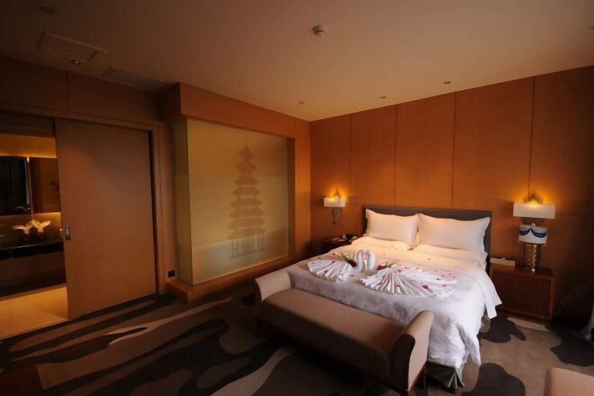 Quarto de hotel com cama, mesas de cabeceira, um banco, porta deslizante e painel decorativo na parede.