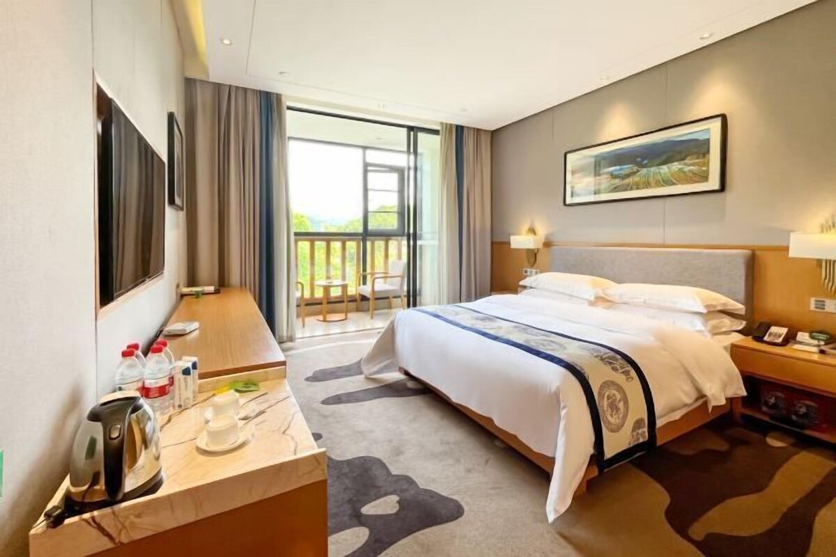 Quarto de hotel com uma cama, uma escrivaninha com chaleira, uma cadeira, varanda com vista e um quadro na parede.
