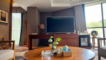 A modern hotel room with a large flat-screen TV, a round wooden coffee table with decorative items, a small cabinet, and a view of the outdoors through large windows.