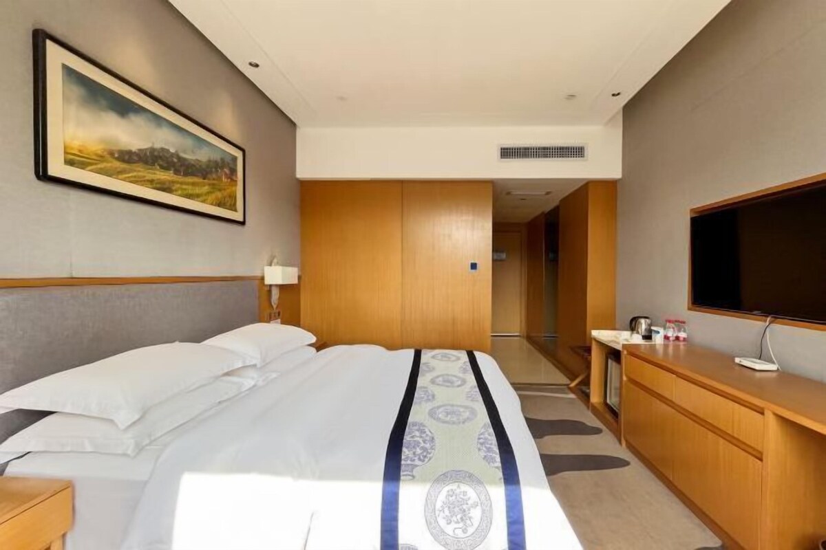 Quarto de hotel com uma cama grande, penteadeira de madeira, televisão e um quadro na parede.