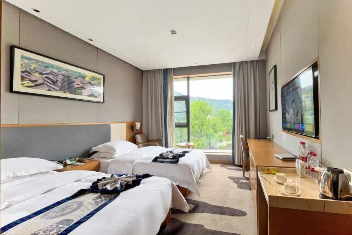 Ein Hotelzimmer mit zwei Betten, einem Schreibtisch mit Fernseher und einem Fenster mit Blick auf Grünflächen.