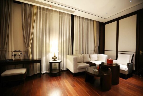 A modern hotel room with a wooden floor, a white sofa, a small coffee table, a side table with a lamp, and a large window with sheer curtains. - Gangcheng Hotel (Zhoushan)