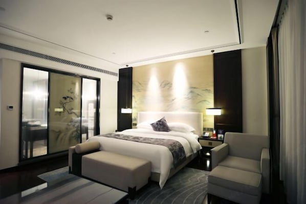 A modern hotel room with a large bed, a sitting area with a chair, a desk, and a large window.