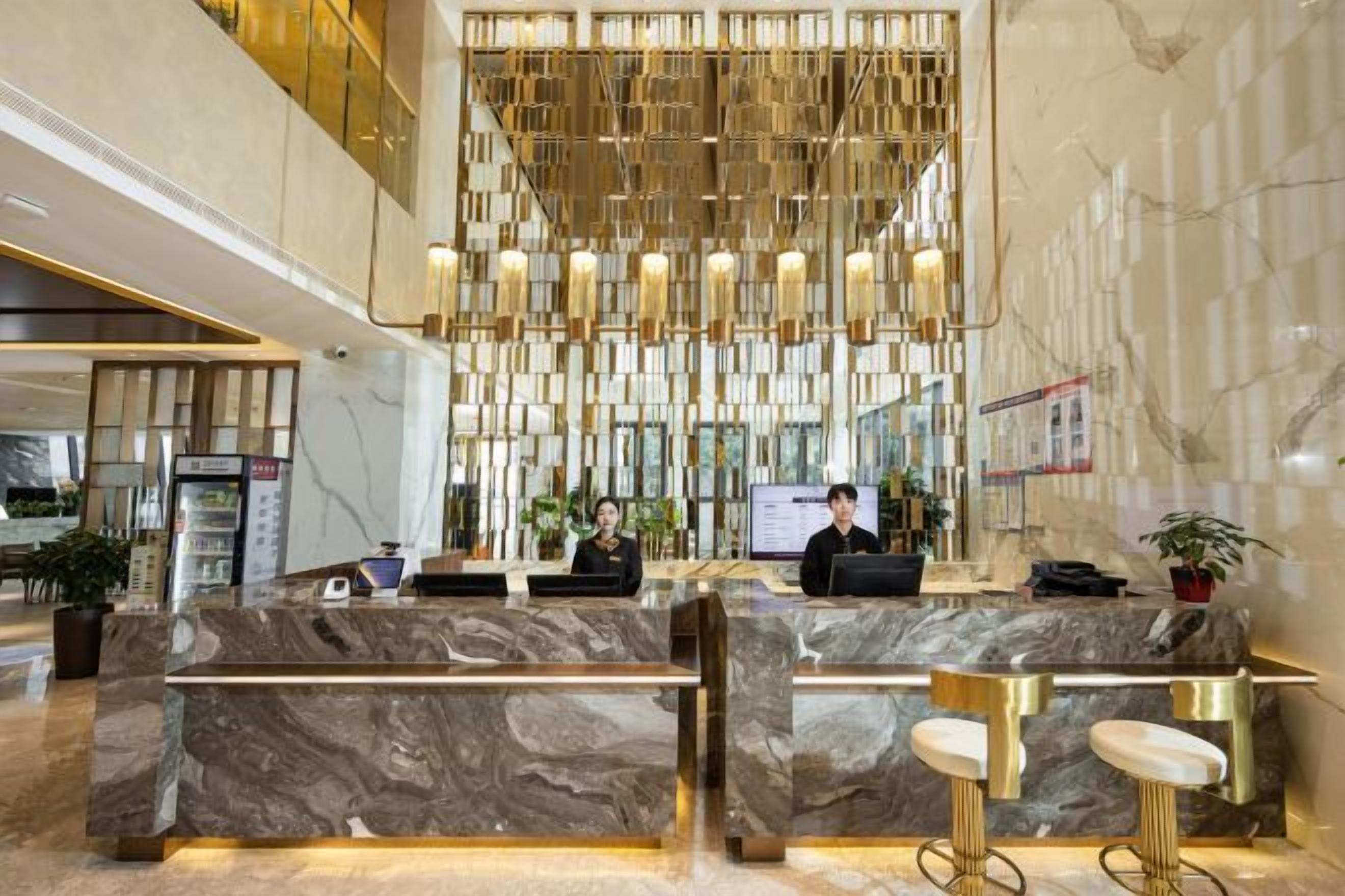 Um hall de hotel moderno com balcões de recepção em mármore, teto alto com luminária geométrica e uma grande tela digital.